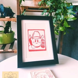 May include: A framed red and white print of a cartoon character wearing a cowboy hat and holding a can. The print is in a black frame, and a carved stamp of the same image is in front of it. The background includes plants and shelves.