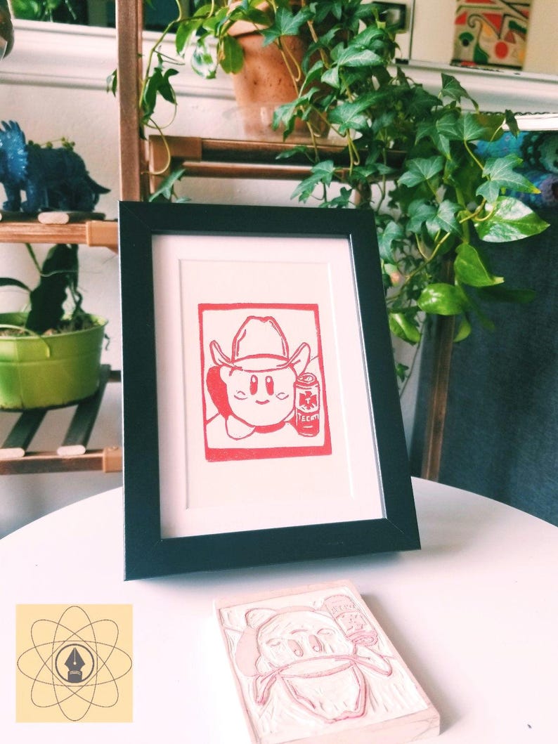 May include: A framed red and white print of a cartoon character wearing a cowboy hat and holding a can. The print is in a black frame, and a carved stamp of the same image is in front of it. The background includes plants and shelves.