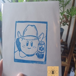 May include: A blue linocut print on white paper features a cartoon character wearing a cowboy hat and holding a can of Tecate beer. The print is framed in a blue rectangle. A small logo is in the bottom right corner.