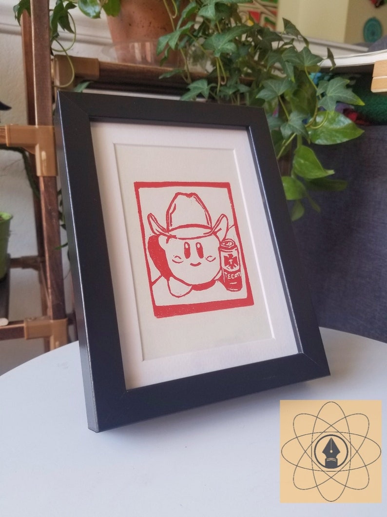 May include: A framed print featuring a red cowboy character holding a can of Tecate beer. The artwork is set against a white mat and framed in a black rectangular frame. The print is displayed on a white surface.