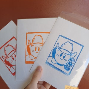 May include: Three art prints featuring a cartoon character wearing a cowboy hat. The prints are in red, orange, and blue ink on white paper. The blue print includes a can of Tecate beer. The prints are held in a hand.