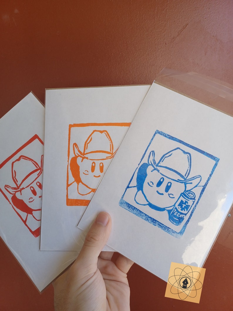 May include: Three art prints featuring a cartoon character wearing a cowboy hat. The prints are in red, orange, and blue ink on white paper. The blue print includes a can of Tecate beer. The prints are held in a hand.