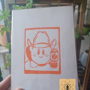 May include: Orange print on white paper featuring a cartoon character wearing a cowboy hat and holding a can of Tecate beer. The image is framed in an orange rectangle. A small logo is in the bottom right corner.