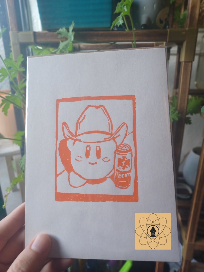 May include: Orange print on white paper featuring a cartoon character wearing a cowboy hat and holding a can of Tecate beer. The image is framed in an orange rectangle. A small logo is in the bottom right corner.