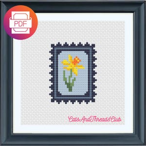 Könnte beinhalten: Kreuzstich-Kunst einer Narzissenblume in einem Briefmarkenrahmen. Die Blume ist gelb und orange mit grünem Stiel und Blättern. Der Rahmen ist dunkelblau und der Hintergrund ist weiß. Der Text "Cats And Threads Club" steht unten.