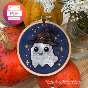 Può includere: Un telaio da ricamo a punto croce con un fantasma sorridente con un cappello da strega, su uno sfondo blu scuro con stelle gialle. Il disegno è incorniciato in un cerchio di legno, con il testo "Cats And Threads Club" in basso.