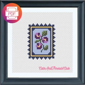 Puede incluir: Patrón de punto de cruz de un diseño floral dentro de un marco de sello postal. El sello presenta flores moradas y hojas verdes sobre un fondo azul claro, enmarcado por un borde azul oscuro. La imagen está sobre un fondo blanco y enmarcada en un marco azul oscuro. El texto "CatsAndThreadsClub" está en la parte inferior.