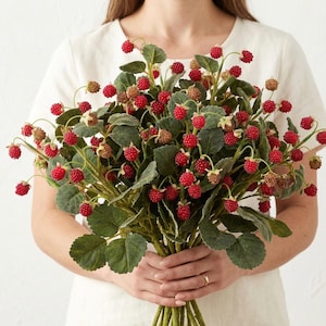 May include: A bouquet of artificial red berries and green leaves. The berries are small and round, with a textured surface. The leaves are a deep green color. The bouquet is held in front of a white background.