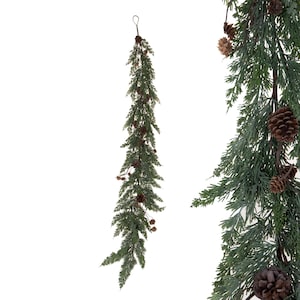 May include: A vertical artificial evergreen swag featuring dark green cedar-like foliage and small brown pinecones. The swag has a thin wire loop at the top for hanging and displays a dense, layered arrangement of needle-like branches.