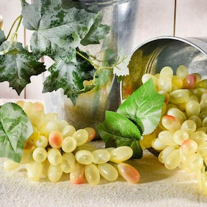 Puede incluir: Dos racimos de uvas artificiales de color verde claro con puntas rosadas y hojas verdes. Un racimo descansa sobre la superficie, mientras que el otro sale de un cubo de metal plateado. Un tallo separado con grandes hojas verdes se mantiene erguido.