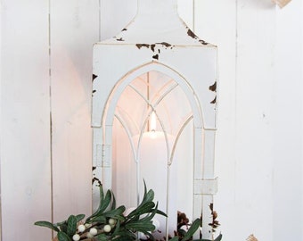 May include: A white distressed lantern with a lit candle inside, decorated with green leaves and white berries. The lantern has an arched window design and sits on a white wooden crate. A burlap ribbon is draped in the background.