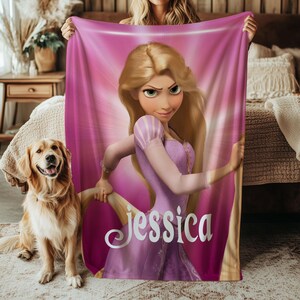 May include: A pink fleece blanket featuring a cartoon princess in a purple dress with the name "Jessica" in white script. The princess has long blonde hair and is set against a pink background. A golden retriever dog is sitting on the left.