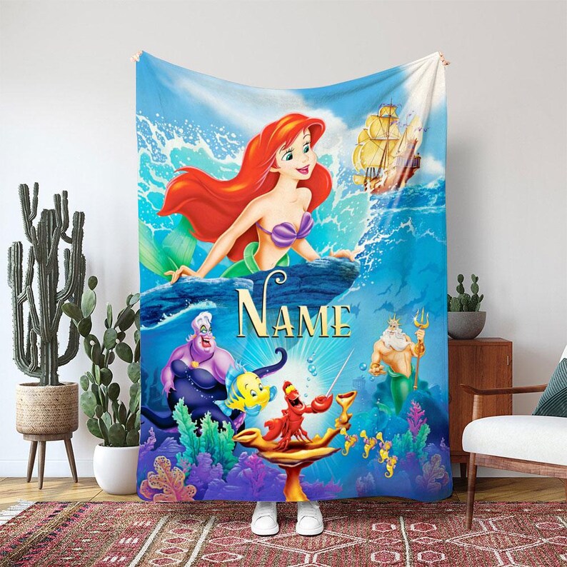 May include: A blue blanket with a vibrant illustration of Ariel from The Little Mermaid. The design includes Ariel, Ursula, Flounder, Sebastian, King Triton, and a ship. The word "Name" is printed in gold. The blanket is displayed indoors.