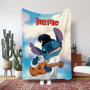 May include: A blue and white blanket featuring a cartoon character playing a ukulele. The character is blue with large ears and is wearing a white outfit. The word "Name" is printed in red at the top.