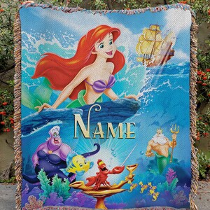 May include: A woven throw blanket with a vibrant illustration of Ariel the mermaid, a ship, and other characters from the animated film. The blanket has a blue background, fringed edges, and the word "NAME" in gold.