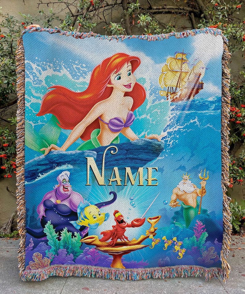 May include: A woven throw blanket with a vibrant illustration of Ariel the mermaid, a ship, and other characters from the animated film. The blanket has a blue background, fringed edges, and the word "NAME" in gold.