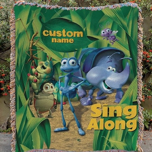 Può includere: Una coperta verde tessuta con personaggi del film d'animazione "A Bug's Life". La coperta ha un bordo con frange e include il testo "custom name" e "Sing Along".