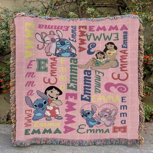May include: Pink woven throw blanket featuring cartoon characters and the name "Emma" in various fonts and colors. The blanket has a fringed edge and a grid pattern. The characters include Stitch and Lilo.