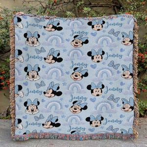 May include: A light blue throw blanket featuring a repeating pattern of Minnie Mouse, bows, rainbows, and stars. The name "Jenny" is written in a script font throughout the design. The blanket has a fringed edge.