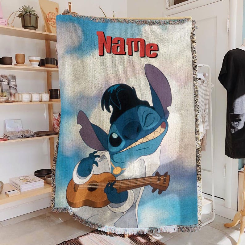May include: Woven throw blanket featuring a blue cartoon character playing a ukulele. The character is wearing a black wig and white outfit. The blanket has a blue and white cloudy background with the word "Name" in red.