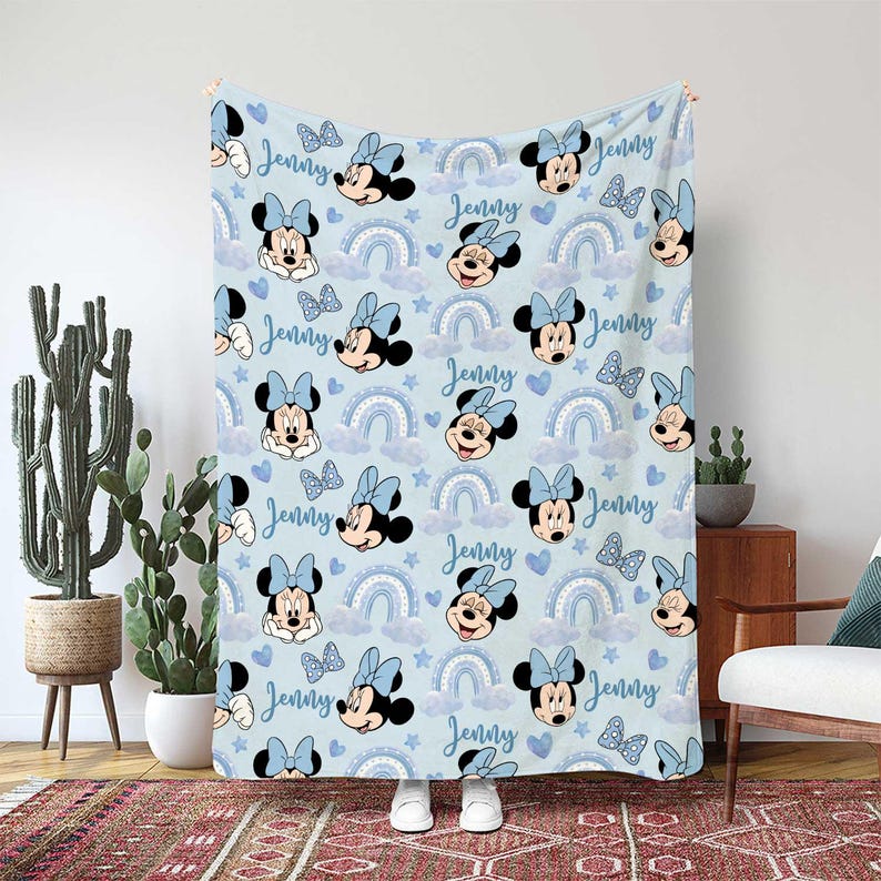 May include: A light blue blanket featuring a repeating pattern of Minnie Mouse, bows, rainbows, clouds, hearts, and stars. The name "Jenny" is written in a script font throughout the design. The blanket is displayed indoors.