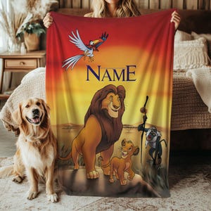 May include: A personalized blanket featuring characters from The Lion King. The design includes Simba, Mufasa, Zazu, Rafiki, and the text "NAME" against a sunset backdrop. A golden retriever sits to the left of the blanket.