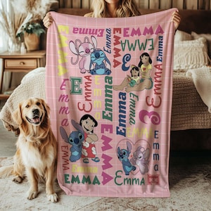 May include: Pink fleece blanket featuring cartoon characters and the name "Emma" in various colors and fonts. The blanket has a grid pattern and includes images of Stitch and Lilo. A golden retriever dog is in the foreground.