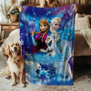 May include: A plush, blue blanket featuring characters from the animated film, Frozen. The blanket showcases Anna, Elsa, and Olaf against a backdrop of snowflakes and a winter landscape. The word "NAME" is visible on the blanket.