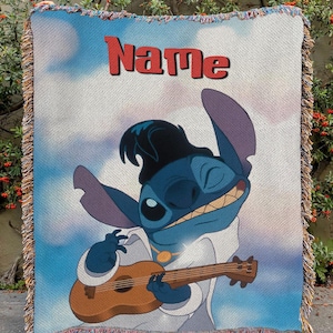 May include: A woven throw blanket featuring a cartoon character playing a ukulele. The character is blue with large ears and is wearing a white outfit and a black hat. The word "Name" is printed in red at the top of the blanket. The blanket has a fringed edge.