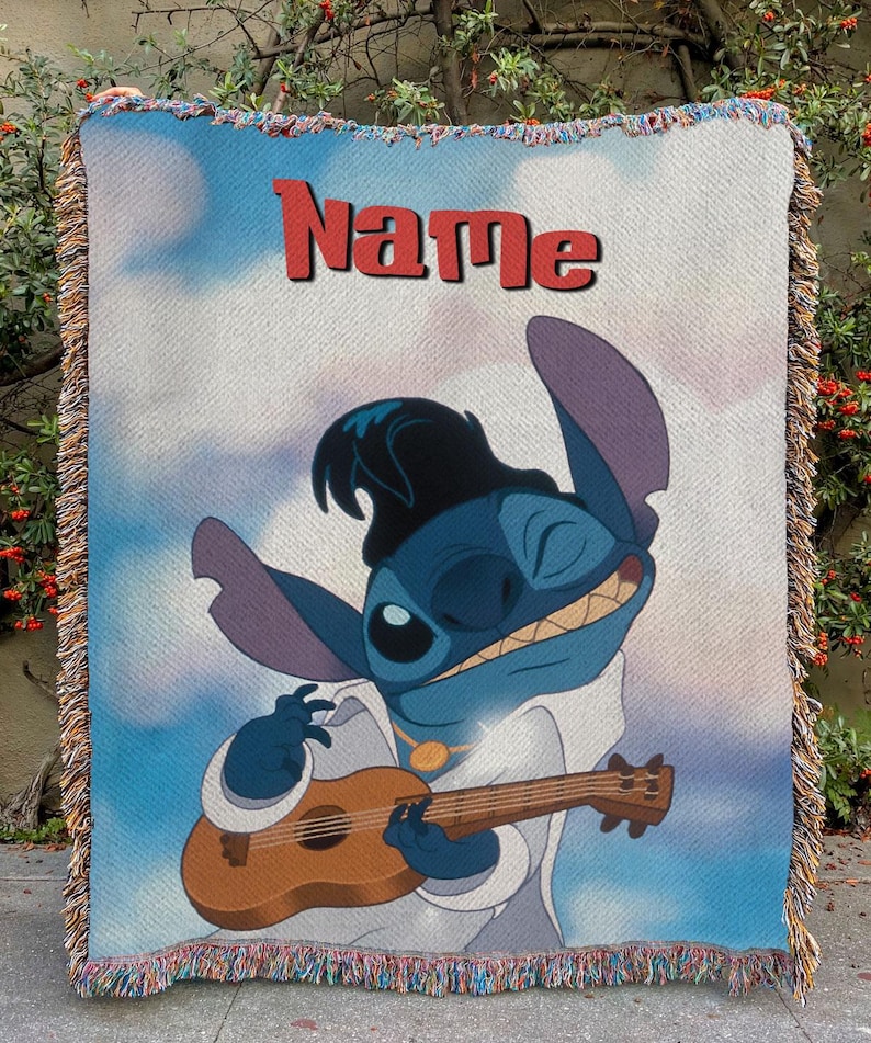 May include: A woven throw blanket featuring a cartoon character playing a ukulele. The character is blue with large ears and is wearing a white outfit and a black hat. The word "Name" is printed in red at the top of the blanket. The blanket has a fringed edge.