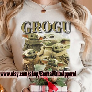 May include: A white sweatshirt with a graphic print of multiple Baby Yoda characters from the Star Wars series. The text "GROGU" is printed in gold above the characters. The text "www.etsy.com/shop/EmmaWhiteApparel" is printed below the characters.