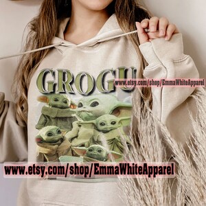 May include: A beige hoodie with a graphic print of multiple green "Grogu" characters from the Star Wars series. The text "Grogu" is printed in green and white letters on the hoodie. The text "www.etsy.com/shop/EmmaWhiteApparel" is printed on the hoodie.
