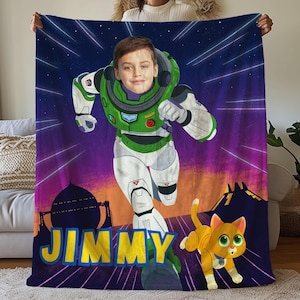 Puede incluir: Una manta de forro polar morada, azul y amarilla con una imagen de dibujos animados de un niño vestido como Buzz Lightyear de la película Toy Story. El niño está corriendo y la manta tiene el nombre "JIMMY" impreso en ella. Un gato amarillo está corriendo detrás del niño.