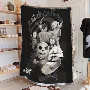 May include: Black and white woven throw blanket featuring characters from The Nightmare Before Christmas. The design includes Jack Skellington, Sally, and other characters, with a graveyard scene and the word "NAME". The blanket has a fringed edge.