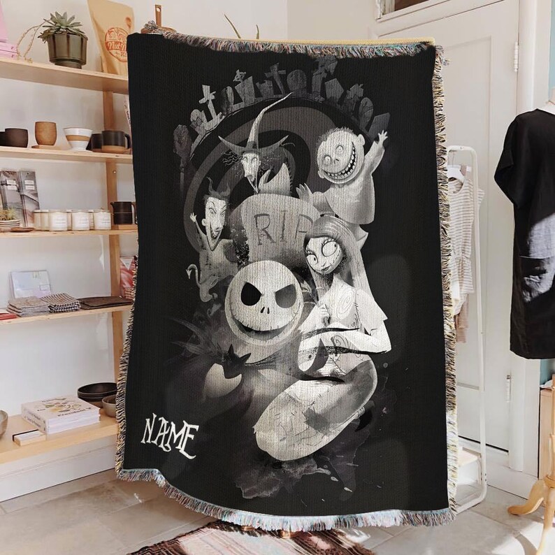 May include: Black and white woven throw blanket featuring characters from The Nightmare Before Christmas. The design includes Jack Skellington, Sally, and other characters, with a graveyard scene and the word "NAME". The blanket has a fringed edge.