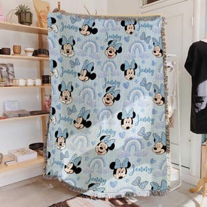 May include: Light blue throw blanket featuring a repeating pattern of Minnie Mouse, bows, rainbows, hearts, and the name "Jenny" in a script font. The blanket has a fringed edge and is displayed indoors.