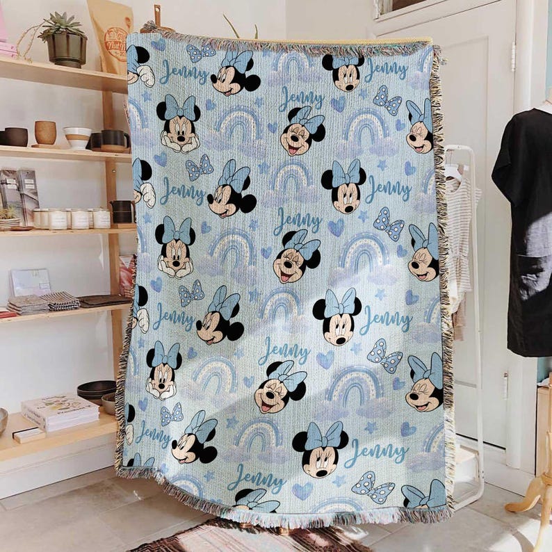 May include: Light blue throw blanket featuring a repeating pattern of Minnie Mouse, bows, rainbows, hearts, and the name "Jenny" in a script font. The blanket has a fringed edge and is displayed indoors.