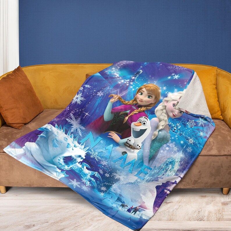 May include: A blue and white Frozen-themed blanket featuring Anna, Elsa, Olaf, and a snow monster. The blanket has a soft, plush texture and is decorated with snowflakes and icy imagery. The blanket is draped over a brown sofa.