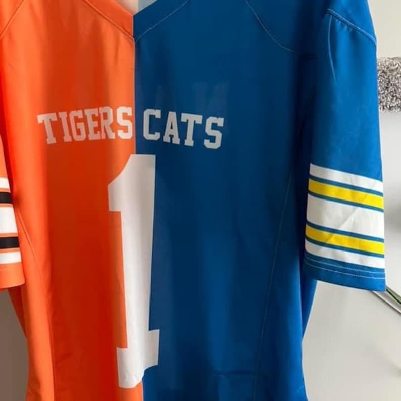 May include: Two football jerseys, one orange with "TIGERS" on the front and one blue with "CATS" on the front. The jerseys are sewn together at the center seam, with a white number "1" on the blue jersey.