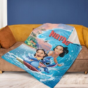 Op de afbeelding: Een blauw-witte deken met een cartoonsc&egrave;ne met de personages Lilo, Stitch en Nani die surfen. De deken heeft een zachte, pluche textuur en het woord "Name" in het rood.