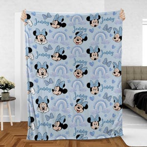 May include: A light blue blanket featuring a repeating pattern of Minnie Mouse, bows, rainbows, clouds, stars, and hearts. The name "Jenny" is written in cursive throughout the design. The blanket is soft and cozy.