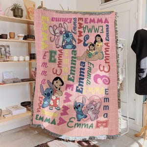 May include: Pink woven throw blanket featuring cartoon characters and the name "Emma" in various colors and fonts. The blanket has a fringed edge and a grid pattern. Characters include Stitch and Lilo.