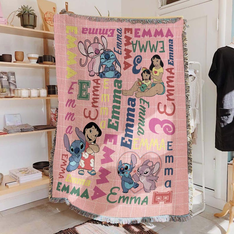May include: Pink woven throw blanket featuring cartoon characters and the name "Emma" in various colors and fonts. The blanket has a fringed edge and a grid pattern. Characters include Stitch and Lilo.