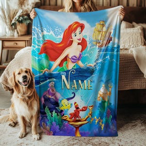 May include: A blue blanket featuring Ariel from The Little Mermaid, with a red-haired mermaid, a ship, and the text "Name". Other characters include Ursula, Flounder, Sebastian, and King Triton. A golden retriever dog is in the foreground.