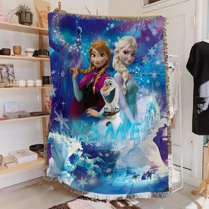 May include: A woven throw blanket featuring characters from the animated film "Frozen." The design includes Anna, Elsa, and Olaf, set against a blue and white winter scene with snowflakes and an ice castle. The word "NAME" is also visible.