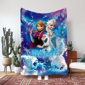 May include: A large, rectangular blanket featuring characters from the animated film Frozen. The blanket displays Anna, Elsa, and Olaf against a blue and purple background with snowflakes and the word "NAME".