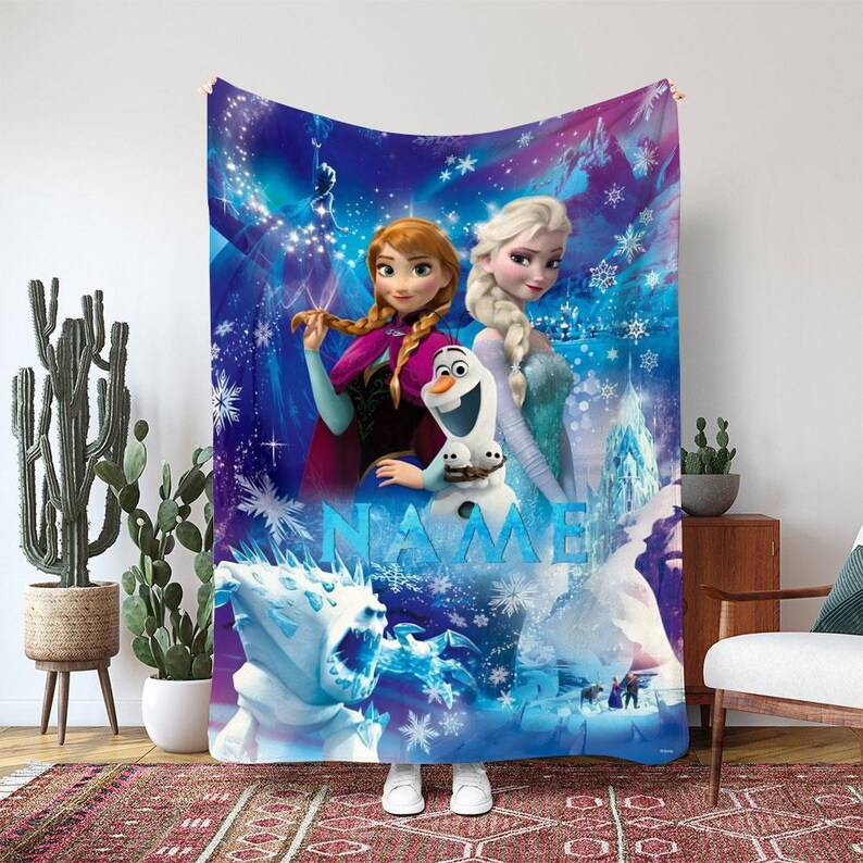 May include: A large, rectangular blanket featuring characters from the animated film Frozen. The blanket displays Anna, Elsa, and Olaf against a blue and purple background with snowflakes and the word "NAME".