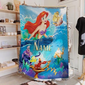 May include: A woven throw blanket with a vibrant illustration of Ariel from The Little Mermaid. The design includes Ariel, Ursula, King Triton, and other characters, with the word "NAME" in gold lettering. The blanket has a fringed edge.