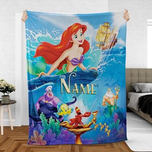May include: A blue blanket featuring Ariel from The Little Mermaid. The blanket has a vibrant illustration of Ariel, Ursula, Flounder, Sebastian, King Triton, and a ship. The word "NAME" is written in gold letters.