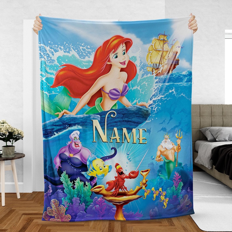May include: A blue blanket featuring Ariel from The Little Mermaid. The blanket has a vibrant illustration of Ariel, Ursula, Flounder, Sebastian, King Triton, and a ship. The word "NAME" is written in gold letters.
